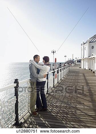 Stock Image - Couple embracing, standing on pier, side view, full length. Fotosearch