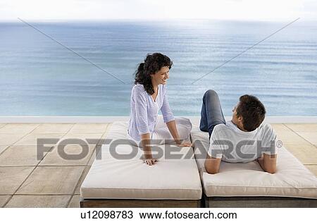 Stock Image - Couple relaxing on sun beds on terrace overlooking sea, elevated view. Fotosearch