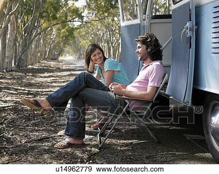 Stock Photo - Couple sitting in deck chairs beside camper van parked at side of road. Fotosearch
