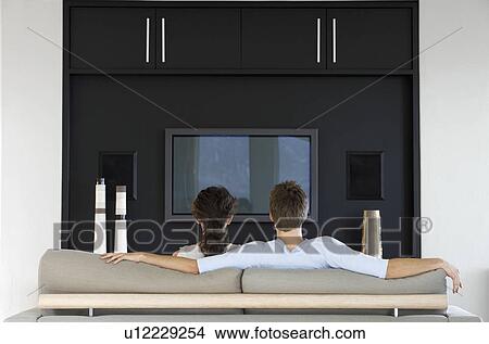 Couple sitting in front of flat screen television in living room, back view View Large Photo Image Picture - Couple sitting in front of flat screen television in living room, back view. Fotosearch