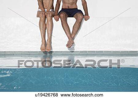 Stock Photo - Couple sitting side by side on edge of swimming pool, low section. Fotosearch