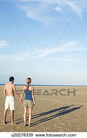 Stock Photo - Couple standing on beach looking out to sea, back view. Fotosearch