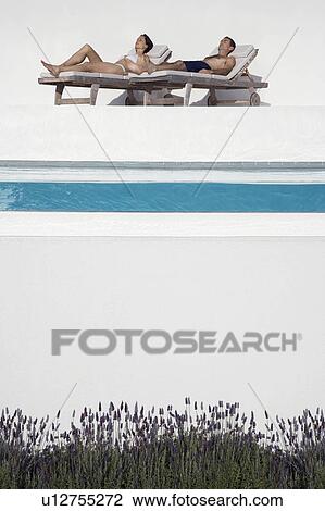 Stock Image - Couple sunbathing on sunloungers by swimming pool. Fotosearch