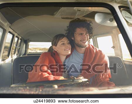 Stock Image - Couple wrapped in blanket in front seat of van, view through windscreen. Fotosearch