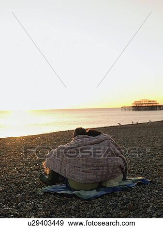 Couple wrapped in blanket sitting on beach, watching sunset, back view View Large Photo Image Stock Photo - Couple wrapped in blanket sitting on beach, watching sunset, back view. Fotosearch