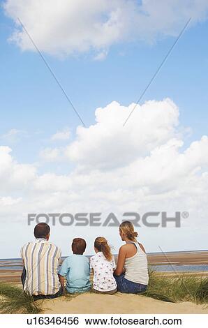 Stock Photograph - Family sitting on sand dune on beach looking at view, back view. Fotosearch