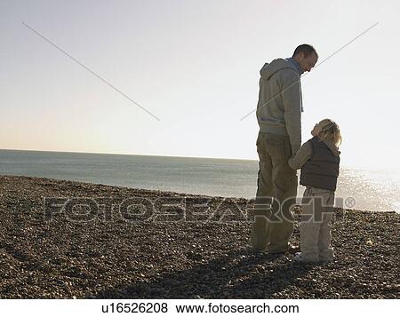 Stock Photo - Father and son looking at each other, standing on beach, full length. Fotosearch