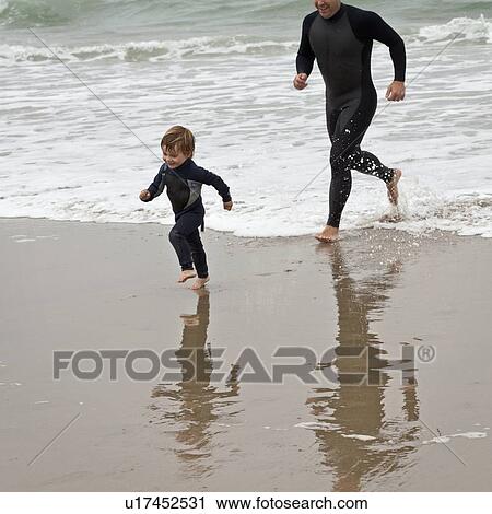 Stock Image - Father and son run barefoot in wetsuits at water's edge. Fotosearch