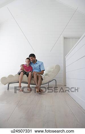 Stock Image - Father and son sitting on lounge chair inside. Fotosearch