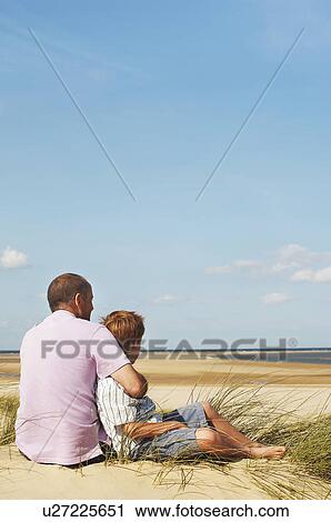 Stock Image - Father and Son sitting on sand dune looking out on Beach, back view. Fotosearch