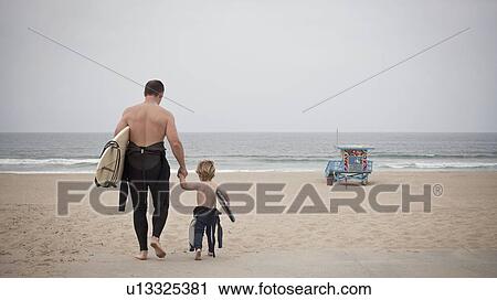 Stock Image - Father and son walk towards sea carrying surfboard. Fotosearch