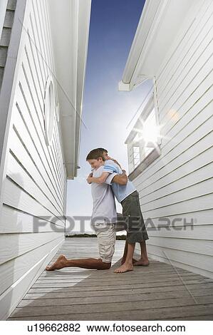 Stock Image - Father hugging son in passageway between buildings, side view. Fotosearch