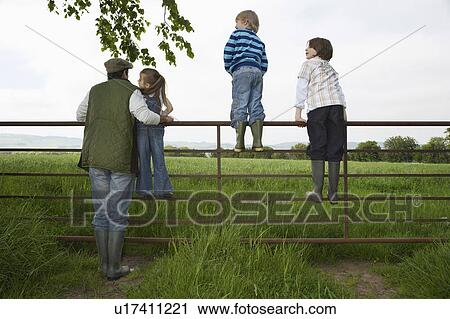 Stock Image - Father with three children (5-9) standing on fence in countryside. Fotosearch