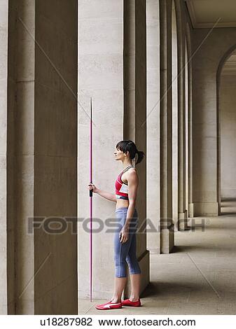 Stock Image - Female athlete holding javelin, standing in portico, side view. Fotosearch