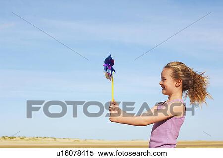 Stock Photography - Girl standing on windy beach holding pinwheel, side view. Fotosearch