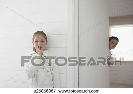 Stock Photo - Girl with finger on lips standing by boy peeking round door. Fotosearch