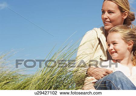 Stock Image - Happy mother and daughter sitting together in long grass against blue sky. Fotosearch
