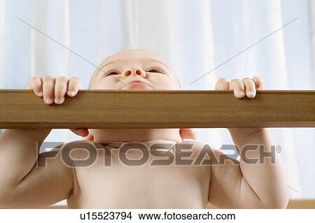 Picture - Infant child holding himself up in wooden cot. Fotosearch