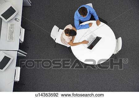 Stock Image - Man and Woman sitting at table using laptop in computer room, overhead view. Fotosearch