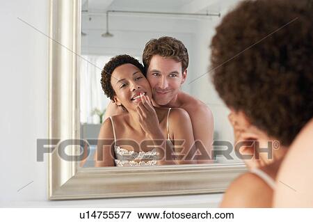 Stock Photo - Man embracing woman applying lipstick in mirror. Fotosearch