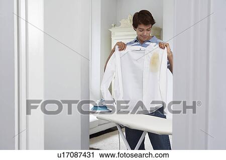 Stock Image - Man holding burned shirt by ironing board. Fotosearch