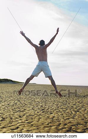Picture - Man on beach doing star jump, back view. Fotosearch