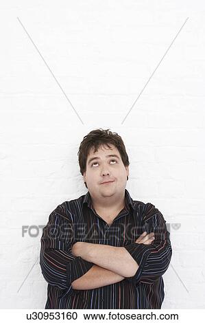 Stock Image - Man standing against wall with Arms Crossed looking upwards, portrait. Fotosearch