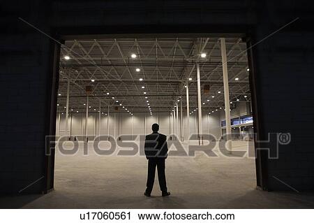 Stock Image - Man stands with hands in pockets at warehouse entrance. Fotosearch