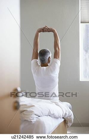 Stock Photography - Man stretching, sitting on edge of bed, back view. Fotosearch