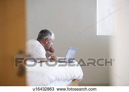 Stock Image - Man using laptop, lying on stomach on bed. Fotosearch