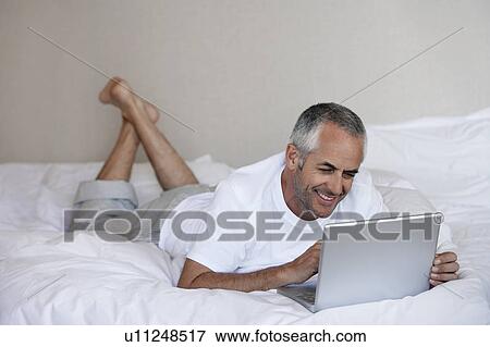 Stock Photo - Man using laptop, lying on stomach on bed. Fotosearch
