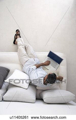 Stock Image - Man using laptop, sitting on sofa in living room, view from above. Fotosearch