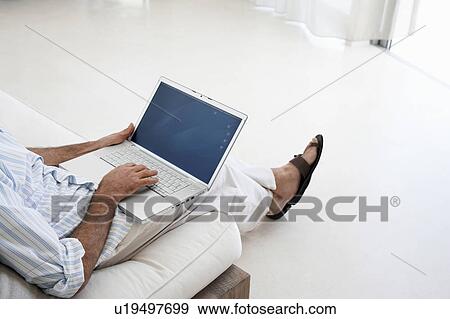 Stock Photo - Man using laptop, sitting on sofa in living room, low section, elevated view. Fotosearch