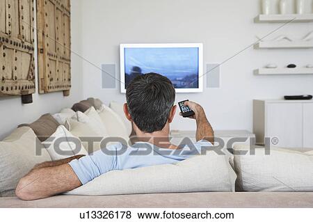 Stock Photo - Man watching television, sitting on sofa in living room, back view. Fotosearch