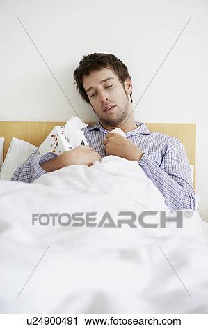 Stock Image - Man with cold in bed holding box of tissues. Fotosearch