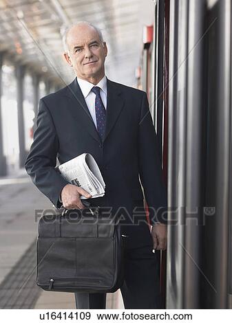 Stock Photo - Mature Businessman holding briefcase and newspaper, standing outside train in empty Train Station. Fotosearch