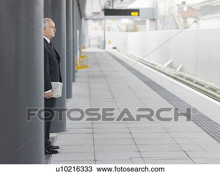 Stock Image - Mature Businessman standing, Waiting at empty Train Station, side view. Fotosearch