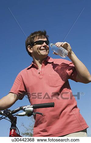 Stock Photo - Mature man stands drinking water and holding bicycle handlebars. Fotosearch