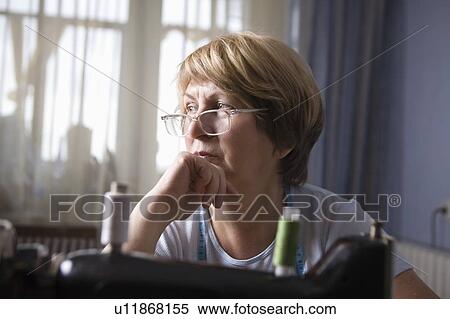 Stock Photography - Mature woman sits leaning on elbow at sewing machine. Fotosearch