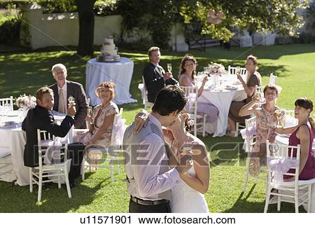 Mid adult bride and groom in garden among wedding guests, holding wineglasses, kissing View Large Photo Image Stock Image - Mid adult bride and groom in garden among wedding guests, holding wineglasses, kissing. Fotosearch