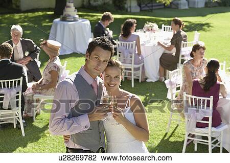 Mid adult bride and groom in garden among wedding guests, holding wineglasses, embracing View Large Photo Image Stock Image - Mid adult bride and groom in garden among wedding guests, holding wineglasses, embracing. Fotosearch