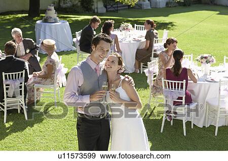 Stock Photo - Mid adult bride and groom in garden among wedding guests, holding wineglasses, embracing. Fotosearch