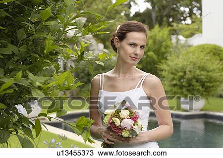 Stock Image - Mid adult bride at poolside, holding bouquet, portrait. Fotosearch
