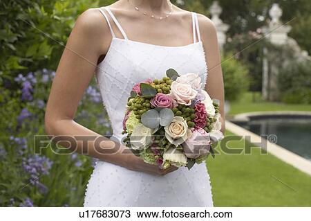 Stock Image - Mid adult bride at poolside, holding bouquet, mid section. Fotosearch