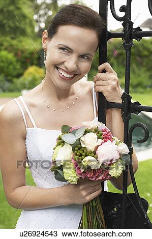 Stock Image - Mid adult bride, leaning on gate, holding bouquet, looking away, portrait. Fotosearch