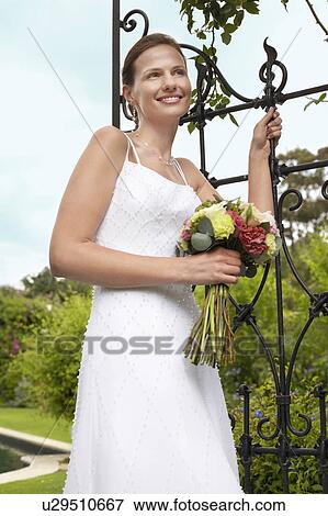 Stock Photo - Mid adult bride leaning on gate, holding bouquet, smiling. Fotosearch