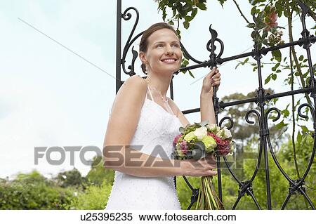 Stock Photography - Mid adult bride leaning on gate, holding bouquet, smiling. Fotosearch