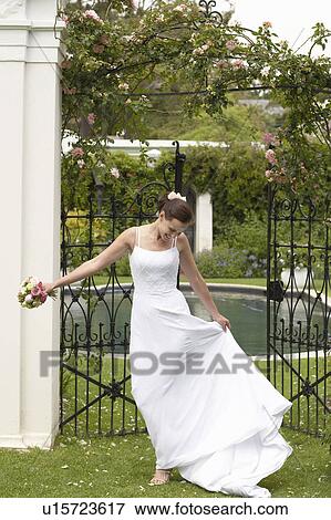 Stock Photo - Mid adult bride standing in garden, holding bouquet and dress, laughing. Fotosearch