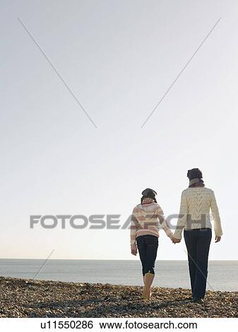 Stock Photograph - Mother and daughter holding hands, walking on beach, back view. Fotosearch