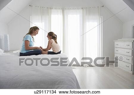 Picture - Mother and daughter sitting on bed in bedroom, side view. Fotosearch